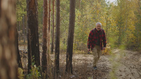 grey-haired-man-with-spin-fishing-rod-in-hand-is-walking-to-shore-of-river-through-forest-area-during-travel