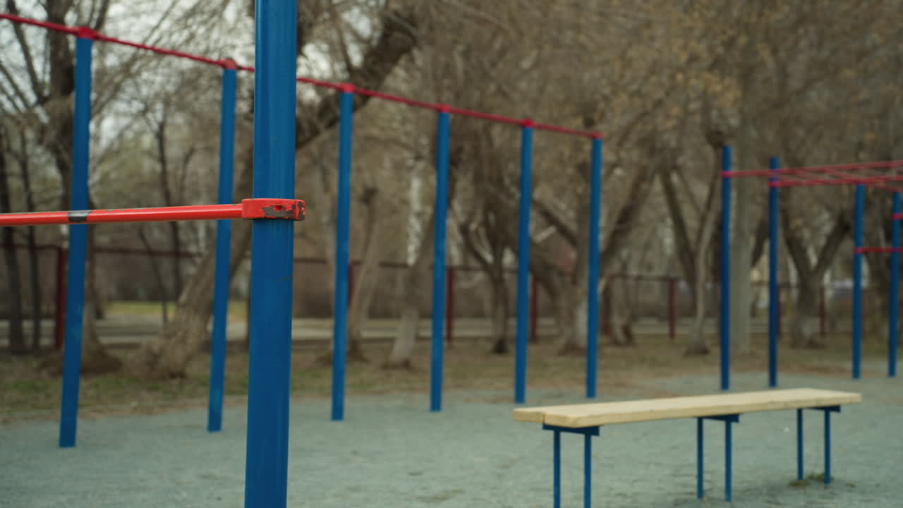 Close-up of blue and red iron bars in a stadium, with two benches positioned in the middle, with barren trees and passing cars visible in the background