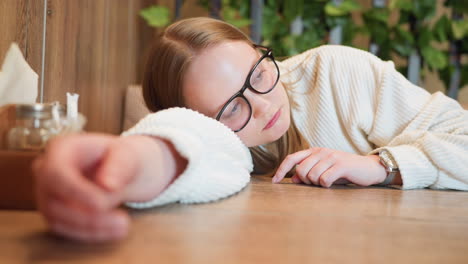 close up of woman resting head on wooden table tapping tabletop lightly with finger wearing glasses and sweater in indoor setting with greenery background