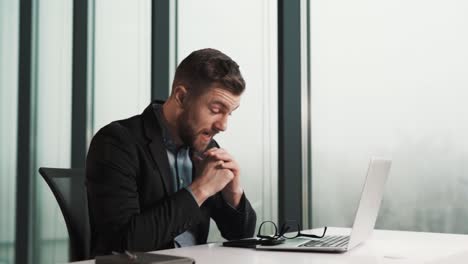 A-worried-man-sits-at-a-table-with-a-laptop-against-the-background-of-panoramic-windows-of-a-skyscraper