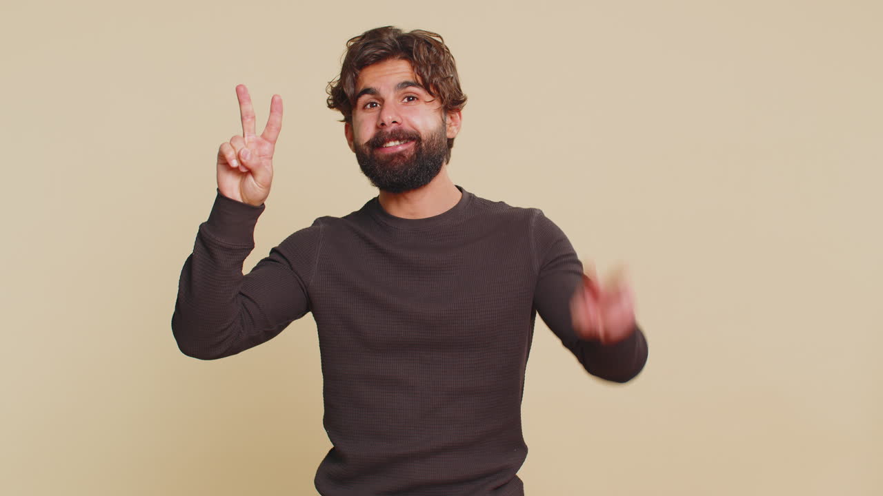 Young indian man showing victory sign doing peace gesture standing against beige studio background