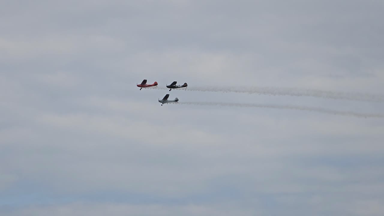 Three vintage aircraft fly in close formation, leaving smoke trails as they soar through a cloudy sky during a flight demonstration.
