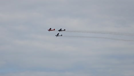 three vintage aircraft fly in close formation, leaving smoke trails as they soar through a cloudy sky during a flight demonstration.