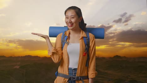 asian female hiker with mountaineering backpack smiling and pointing to side while standing on the top of mountain during sunset time