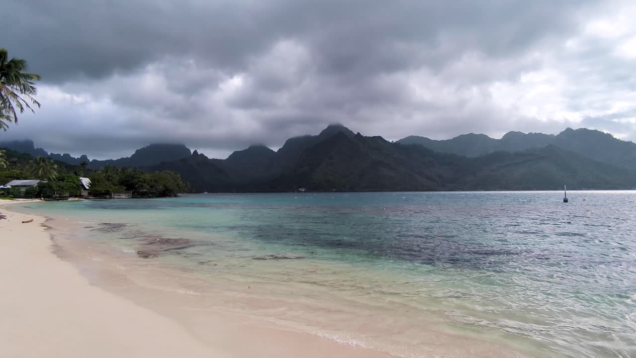 Scenic coastal view of white sandy beach and crystal clear ocean water with rain storm clouds on tropical island of Moorea, French Polynesia