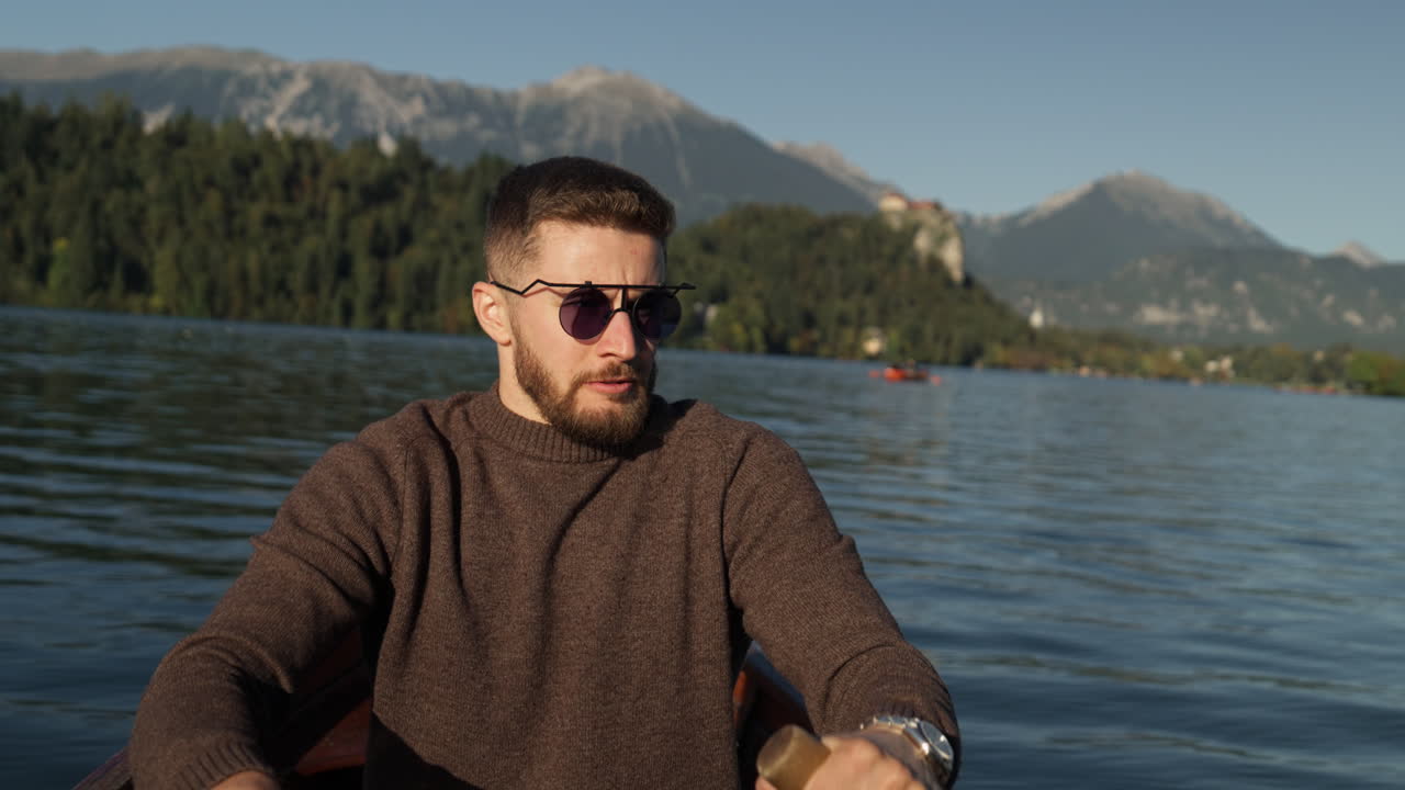A man rowing a wooden boat on a calm lake during autumn, surrounded by vibrant foliage and sunny weather in a serene setting