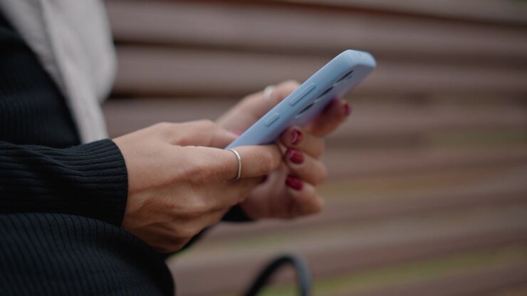Primo piano delle mani che tengono uno smartphone azzurro mentre i pollici toccano, scorrono e navigano durante la navigazione casuale e la messaggistica su una panchina di legno all'aperto con una luce naturale soffusa e uno sfondo di doghe sfocate.