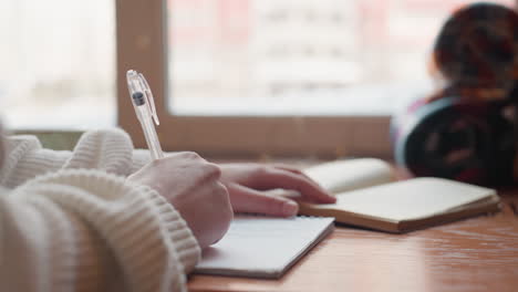 close up hand view of lady in fluffy sweater writing with transparent pen on notepad near open book on wooden table beside window in warm daylight creating calm creative atmosphere for studying