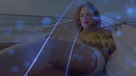 Animation-of-blue-spots-and-light-trails-over-caucasian-woman-using-digital-tablet-on-a-hammock