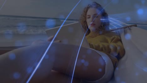 animación de manchas azules y rastros de luz sobre mujer caucásica usando tableta digital en una hamaca