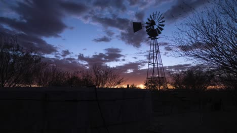 Timelapse-De-Una-Bomba-De-Viento-Y-Un-Tanque-De-Agua-Frente-A-Una-Espectacular-Puesta-De-Sol-Con-Nubes-Anaranjadas-Que-Se-Convierten-En-Estrellas-En-Una-Granja-De-Ovejas-Cerca-De-Keetmanshoop,-Namibia