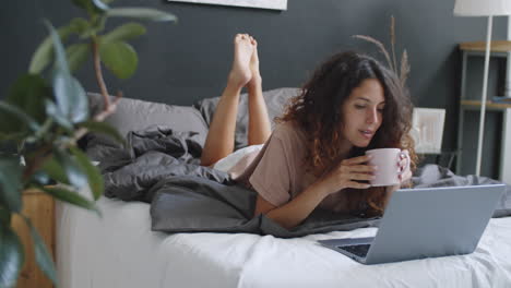 mujer relajándose en la cama con una computadora portátil y café