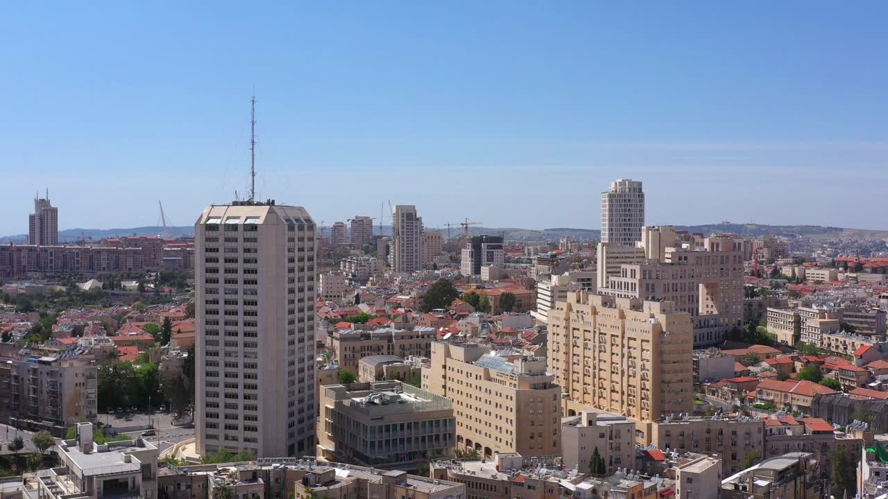 Aerial view of a bustling city skyline featuring various architectural styles, tall buildings, and traditional red-tiled rooftops under a clear blue sky, likely in Jerusalem