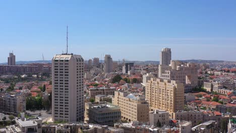 aerial view of a bustling city skyline featuring various architectural styles, tall buildings, and traditional red-tiled rooftops under a clear blue sky, likely in jerusalem