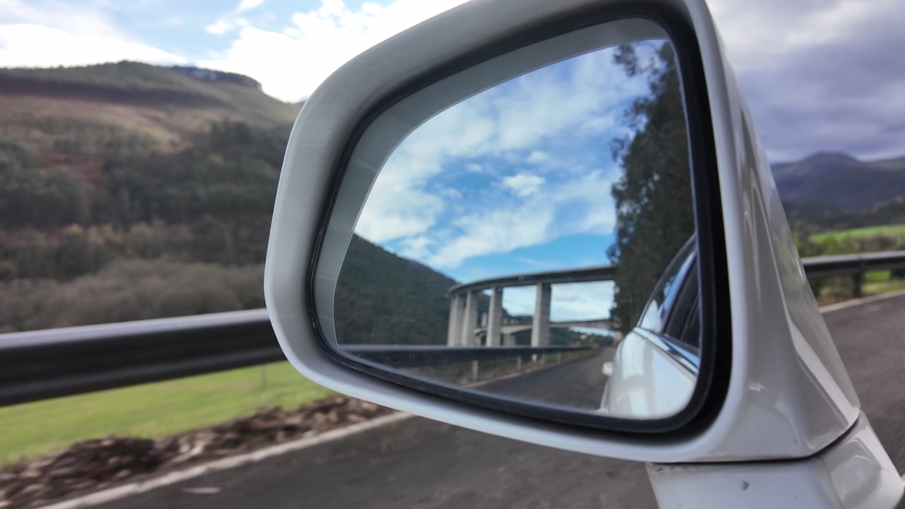 Concrete highway bridge appears reflected in a car rearview mirror while driving beneath cloudy skies, filmed in slow motion.