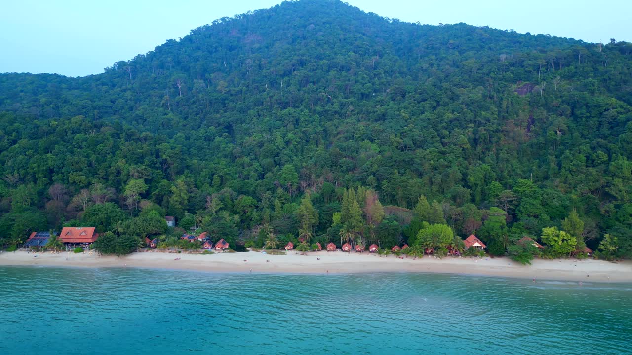 Koh Chang Tropical beach with bungalows along the shore and dense green jungle covering a mountain. Great aerial view descending drone