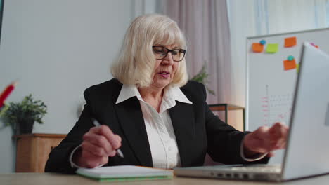 senior elderly business office woman elearning online writing notes from laptop, live webinar online