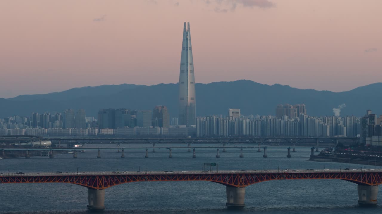 Splendida inquadratura ravvicinata da drone della Lotte World Tower con montagne in controluce e cielo rosa al tramonto, traffico sul ponte di Seongsu e ponti sul fiume Han in primo piano