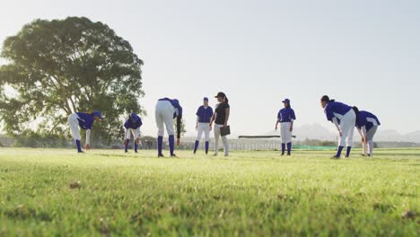 diverse group of female baseball players and coach on pitch, warming up, touching toes