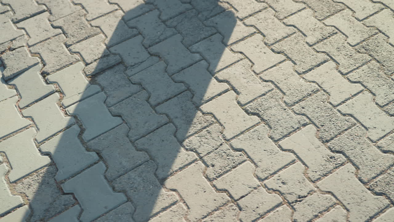 Long shadow across patterned pavement pink coat and boots, silhouette stretching over manhole cover, contemplative commuter slowing step, cinematic angle emphasizing geometry and tile texture, quiet