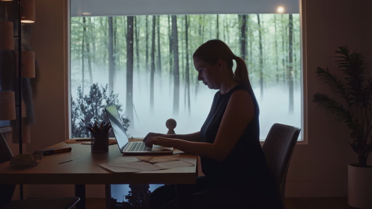 Woman working on a laptop in a home office with a view of a misty forest