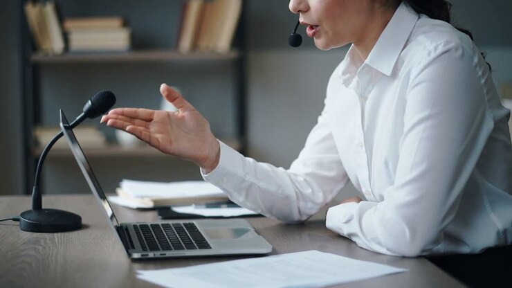 Una donna in camicia bianca che partecipa a una chiamata, presenta e gesticola verso il laptop alla scrivania, con cuffie e microfono.