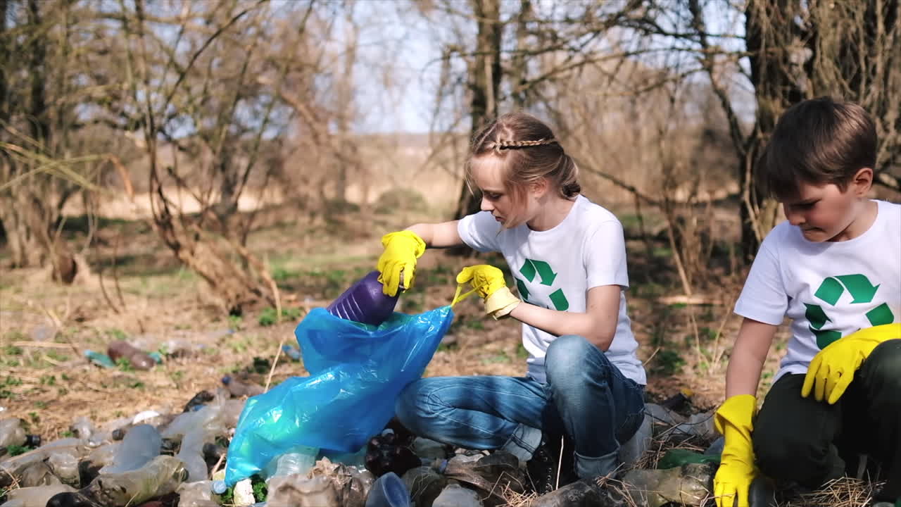 Ragazzo e ragazza che raccolgono rifiuti di plastica in un sacco in uno spiazzo inquinato, simboli di riciclaggio sulle magliette. Rallentatore