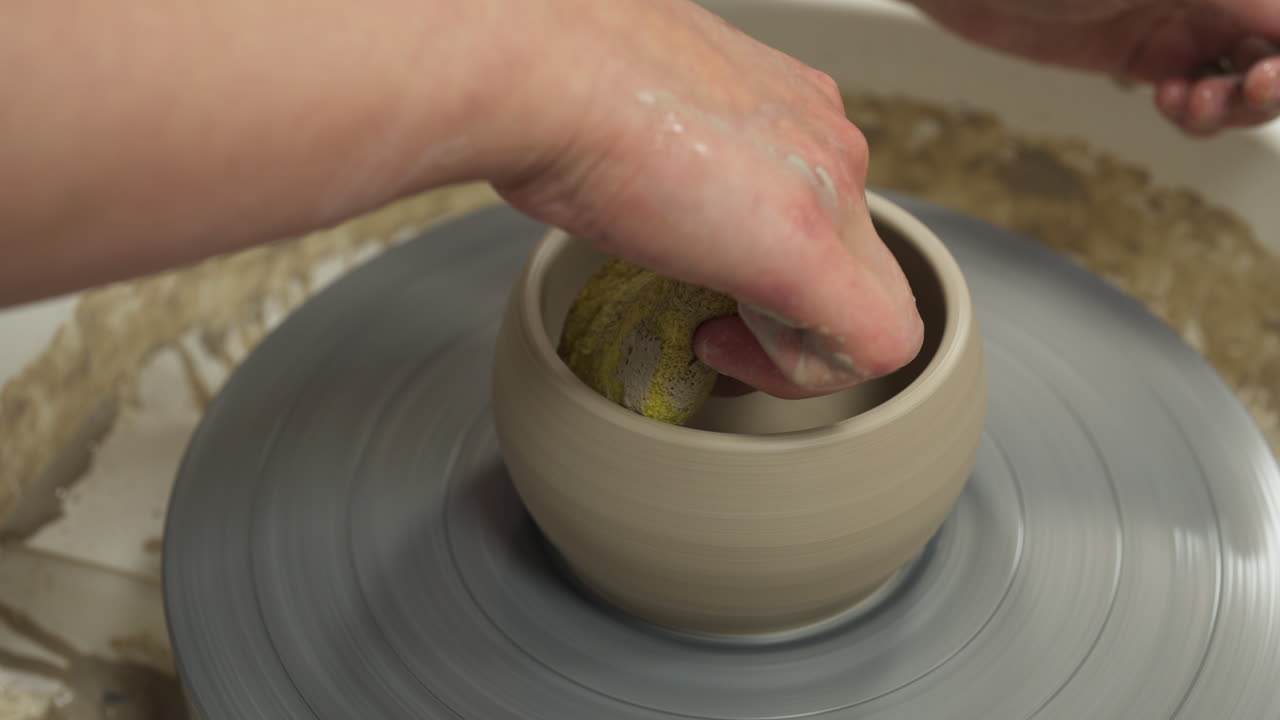 Medium shot of white hands making mug from clay with a yellow sponge, interior