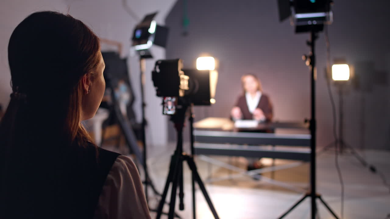 Rear view of a long-haired girl standing at backstage in the photo studio. Lady looks at camera display filming a lady sitting at desk. Blurred backdrop.