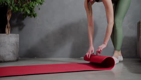 close up a happy brunette girl in a sports summer uniform unrolls a red sports mat and steps on it before starting her sports activities in a modern apartment at home