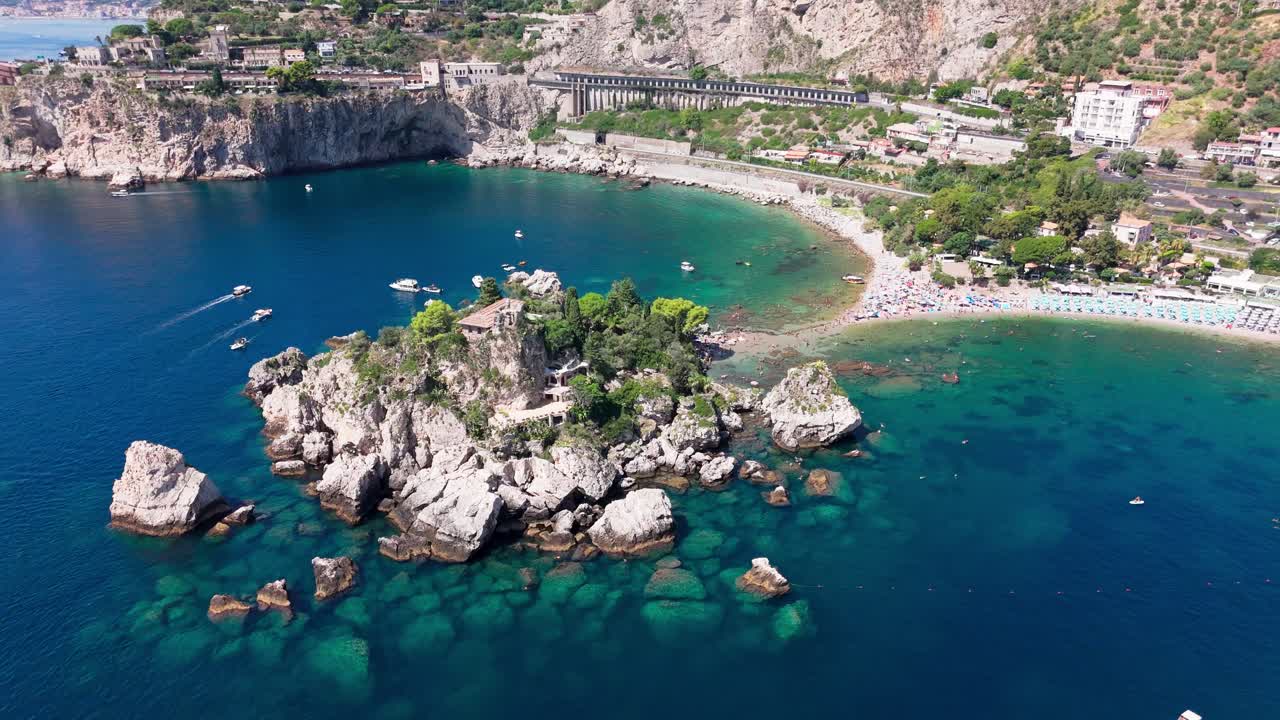 Drone orbits closer from right to left, highlighting Isola Bella, the beach, and detailed coastal features with clear water and rocks