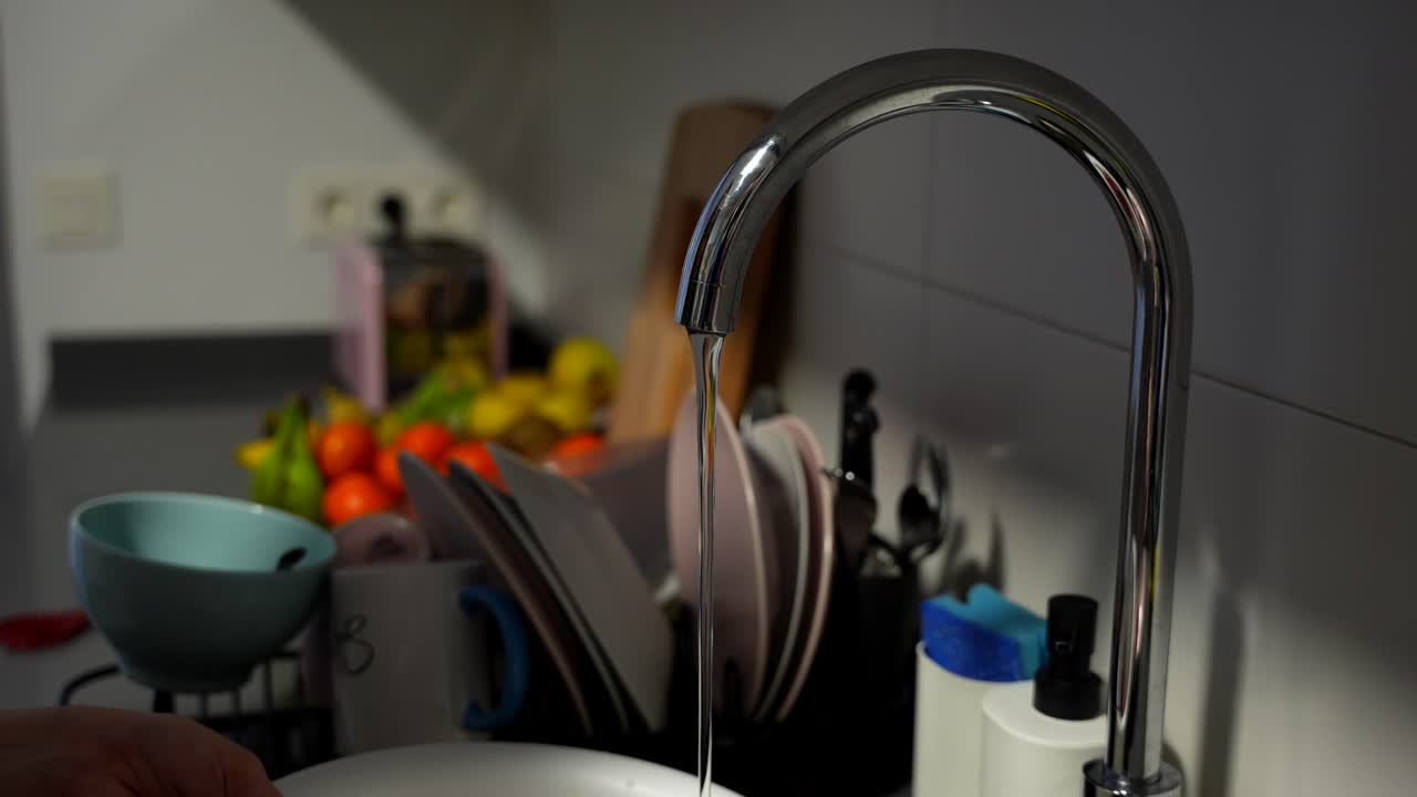 Person washes dishes under running water in kitchen sink, water waste concept