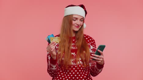 girl in christmas sweater using credit bank card, smartphone, transferring money shopping online