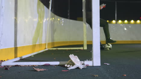 close-up of goalkeeper gloves and scattered leaves on ground blown by the wind near goalpost at night, illuminated by urban lights in background with blur view of someone resting on the goalpost