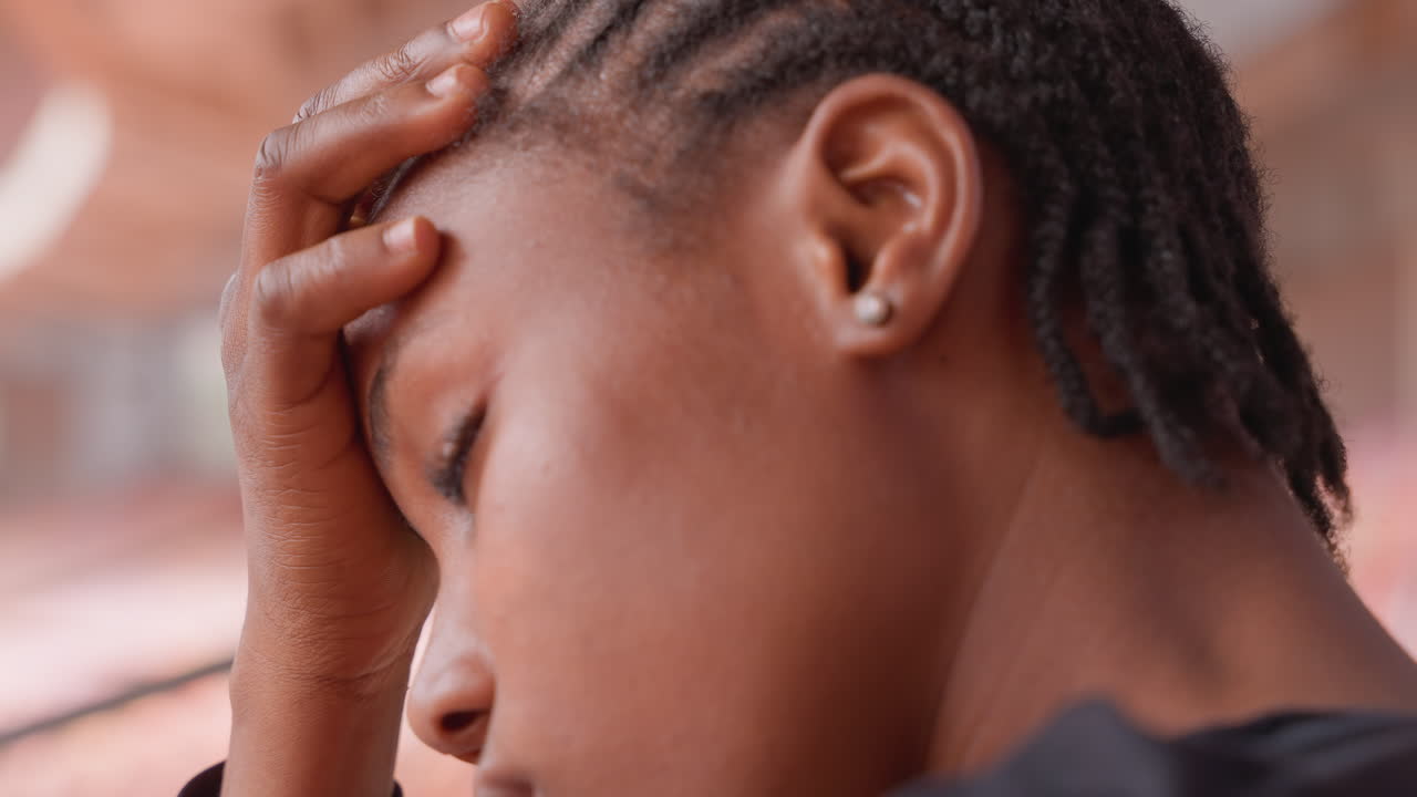 Ragazza adolescente di colore stanca che si tiene la fronte, ritratto ravvicinato di studentessa sopraffatta con occhi chiusi e leggero sudore, capelli intrecciati che incorniciano il viso, sfondo urbano sfocato, emozione pensierosa per lo stato mentale.