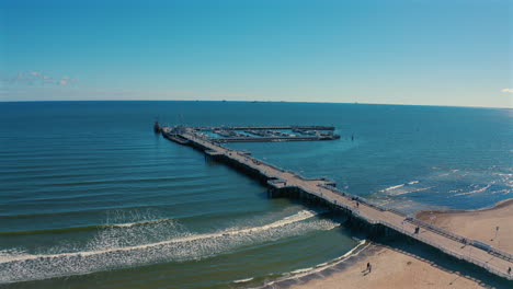 Drone-flying-towards-the-pier-with-ocean-waves-coming-to-the-beach-and-sky-in-the-background