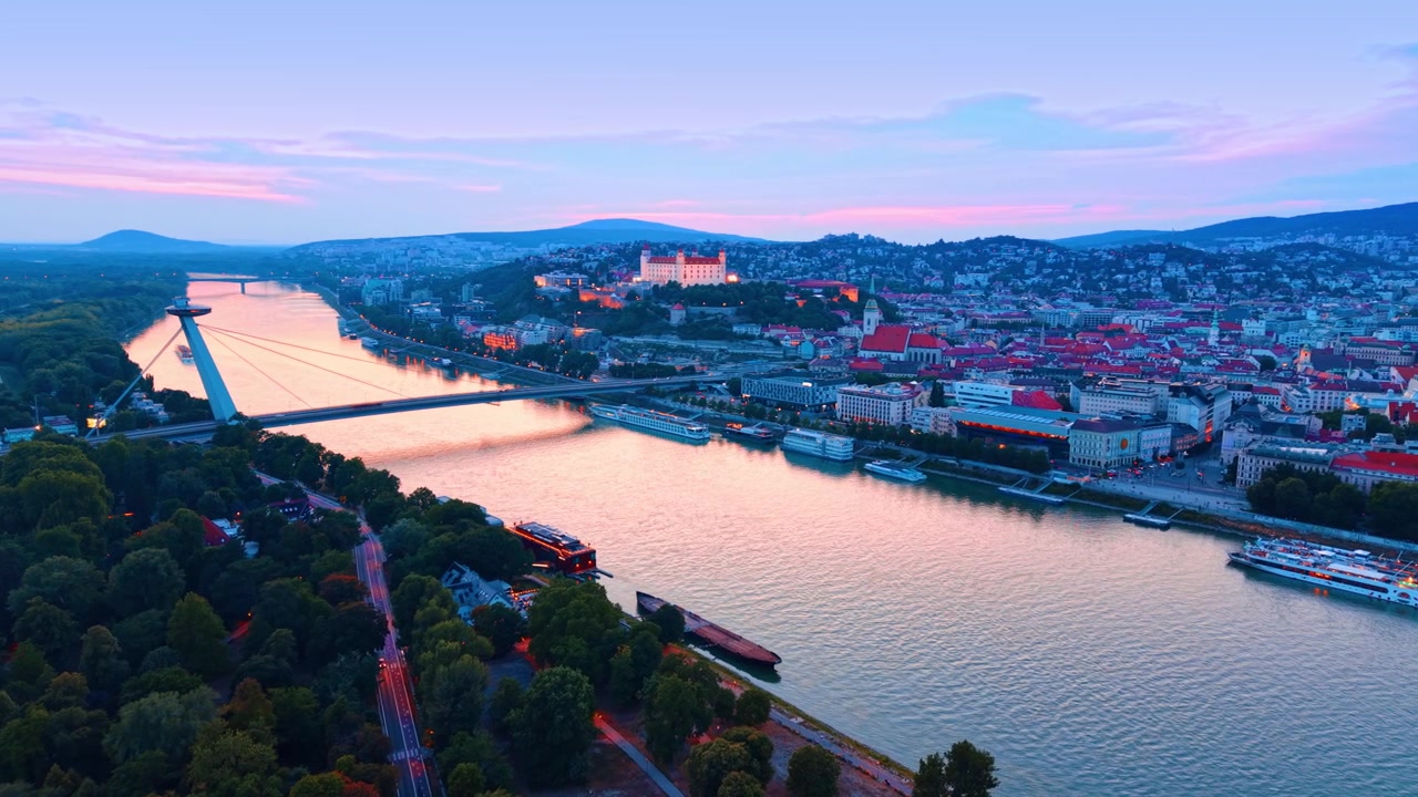 Luftfoto af Bratislavas bybillede og Donau-floden i skumringen. Fugleperspektiv af floden, grønne parktræer, oplyst slot og bycentrum under en lyserød himmel.