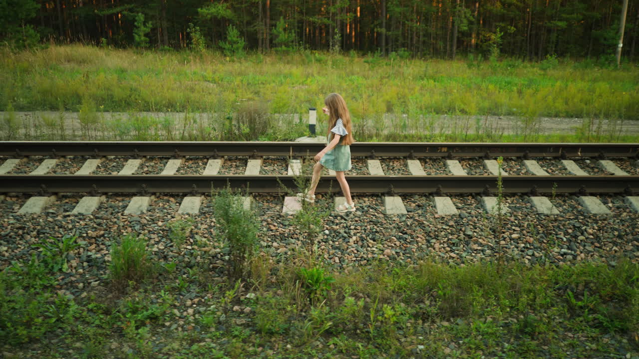 Kid trying to maintain balance on railway sleeper while walking across track, dressed in denim dress and striped shirt, surrounded by gravel, wild plants, and forest in peaceful rural environment