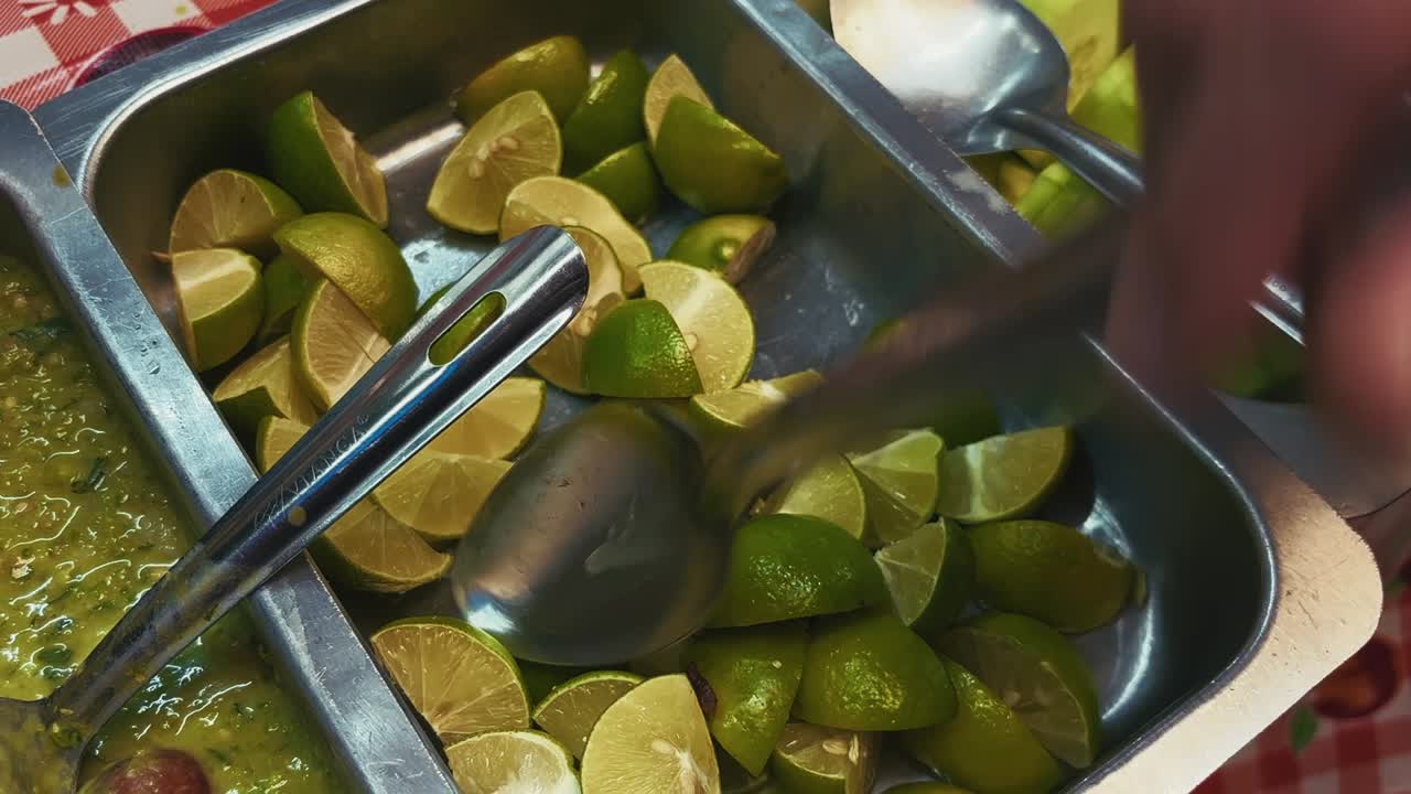 Close-up of fresh lime wedges, sliced cucumbers, and green salsa in metal trays, ready to accompany tacos or other Mexican street foods