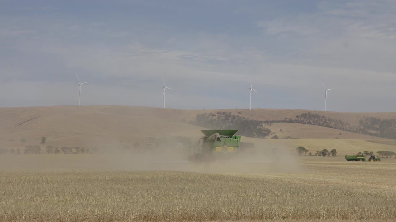 Static shot showing fields and a combine harvester raising a dust cloud with hills and wind turbines in the background