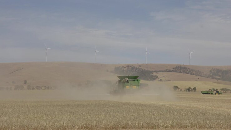 Static shot showing fields and a combine harvester raising a dust cloud with hills and wind turbines in the background