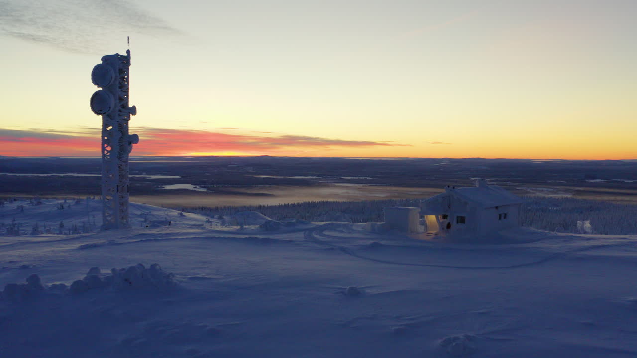 Aerial view reversing across snow covered Lapland remote cabin and communications tower overlooking polar wilderness at sunrise