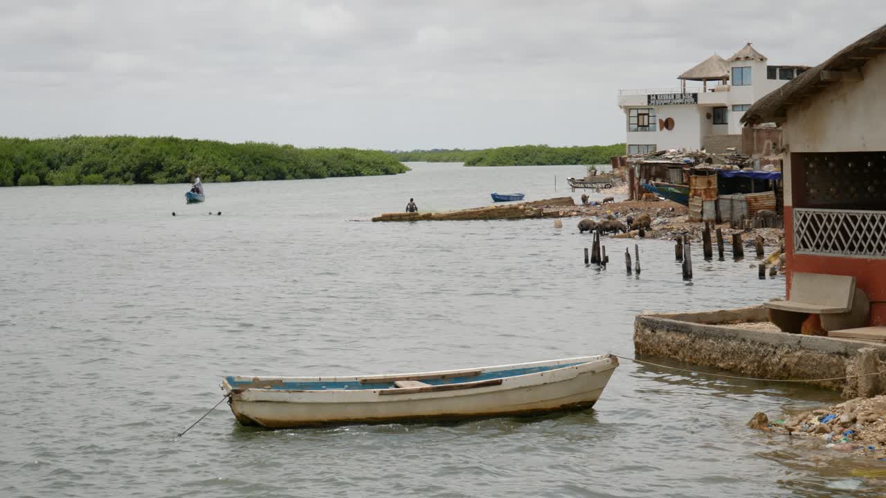 una toma de trípode de la costa en la isla fadiouth, con un barco anclado en primer plano, con casas y algunos cerdos en el fondo