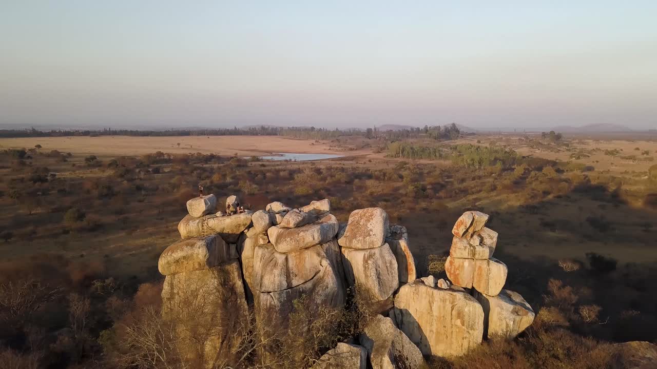 Group of people stand on top of a large mound of boulders, giving them an incredible panoramic view of the surrounding african landscape as the sun goes down.