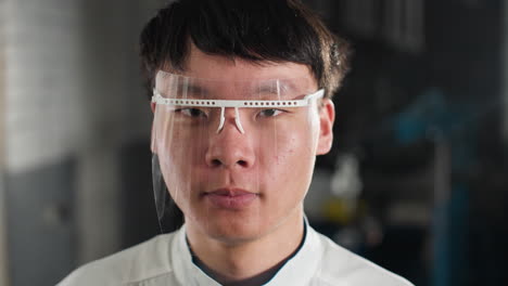 close-up of lab technician wearing white lab coat and protective face shield, blinking with a bold expression, industrial setting with blurred background, soft lighting