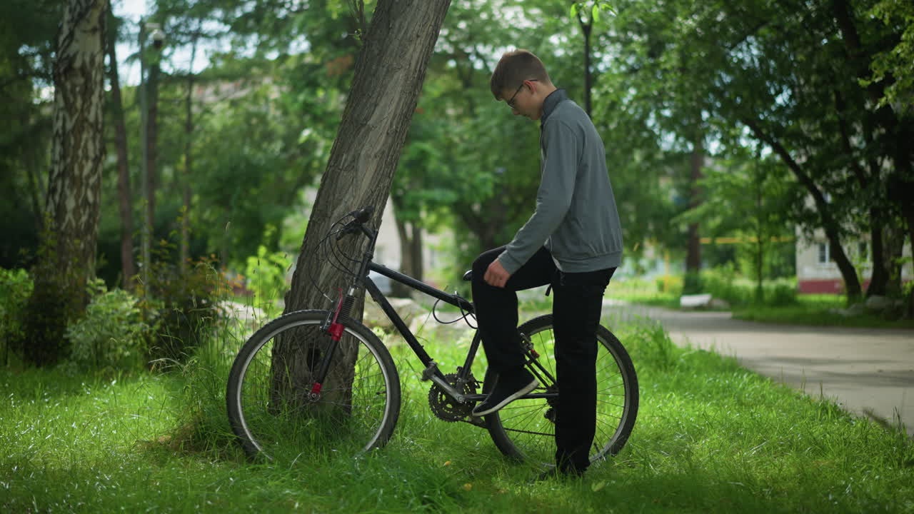 소년은 잔디에 있는 나무 근처에 주차된 자전거를 향해 걸어가고, 그는 페달을 누르고 자전거에 발을 고, 배경에는 소년과 소녀가 배경에서 지나간다.