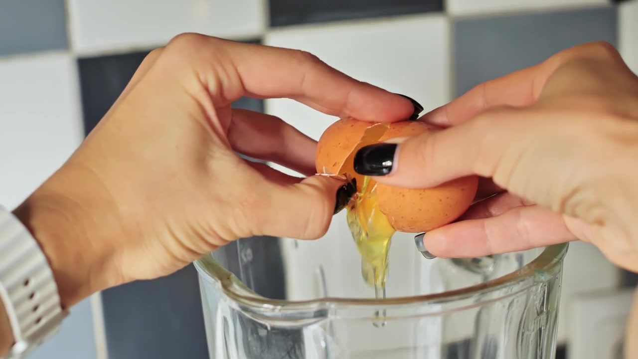 Primer plano de las manos de una mujer rompiendo un huevo en una licuadora para cocinar.