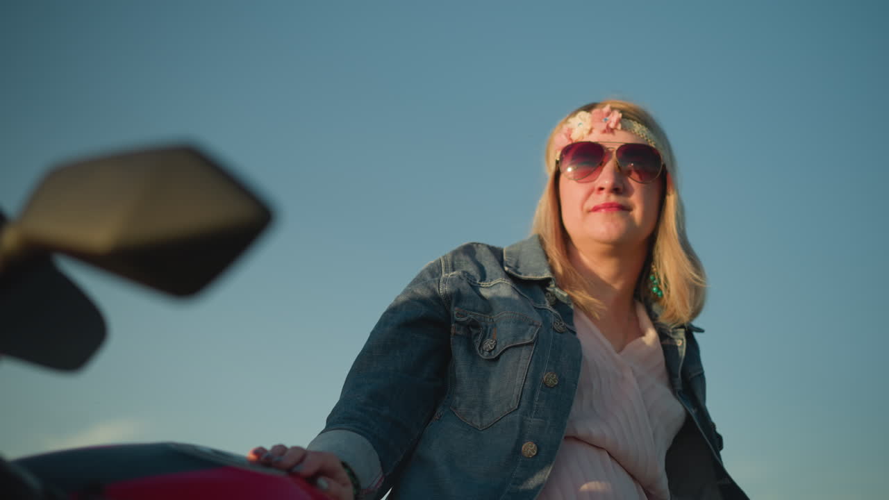 una mujer de mediana edad con una correa de flores y gafas de sol se apoya contra una motocicleta mientras mira pensativamente a la distancia, está vestida casualmente con una chaqueta de vaqueros