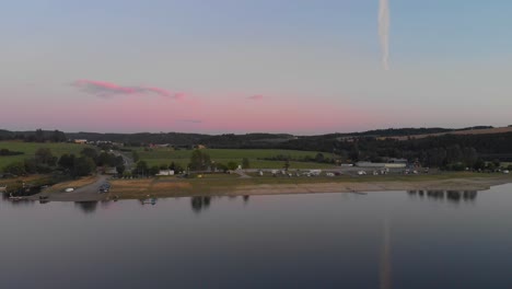 Un-Vuelo-Aéreo-De-Aspecto-Profesional-Hacia-Un-Campamento-Alemán-En-Un-Tranquilo-Lago-Especular-Mientras-El-Cálido-Atardecer