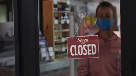 caucasian female shopkeeper wearing face mask changing closed to open sign in window of sports shop